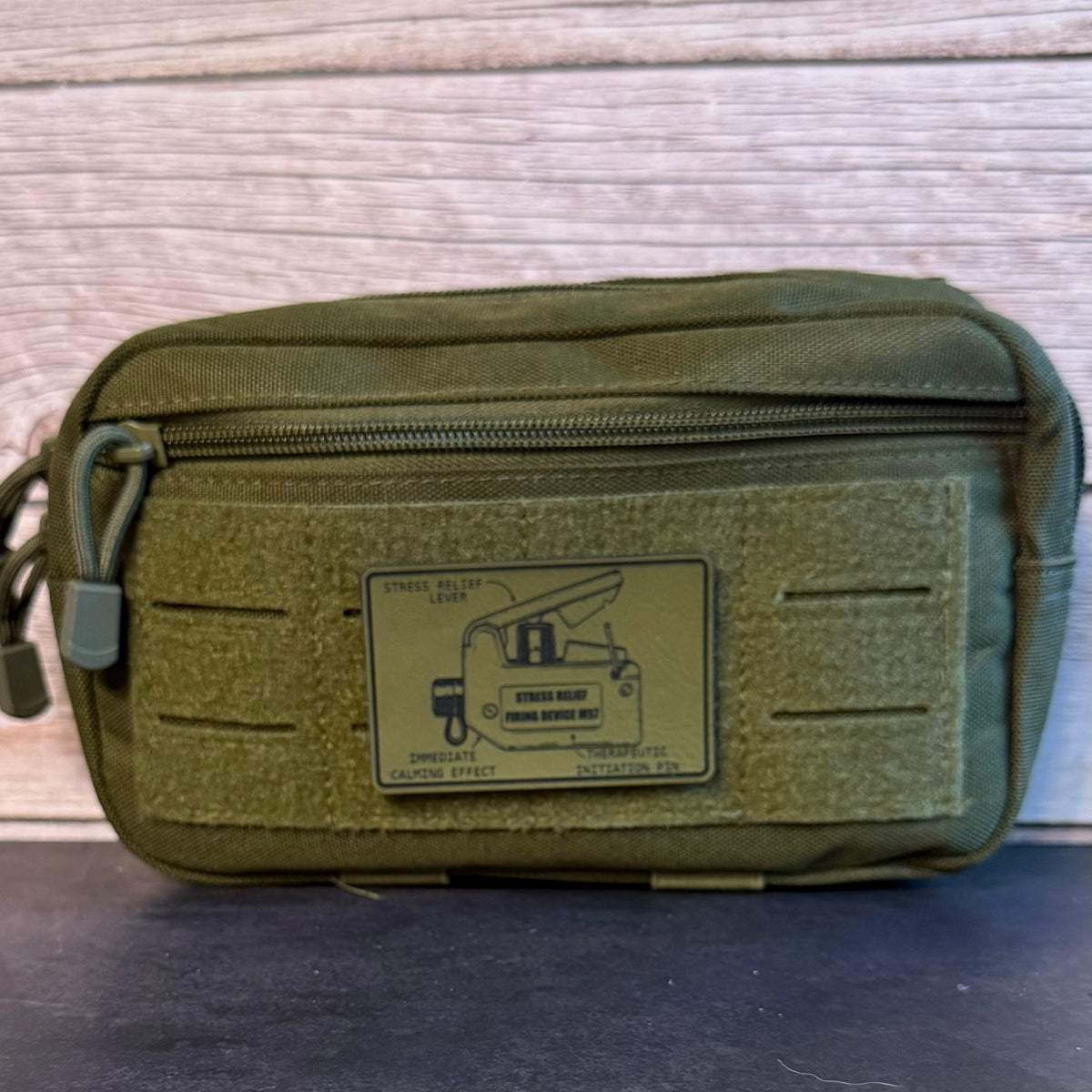 An olive green tactical pouch with a top zipper and a rectangular patch on the front, photographed against a light wood background.