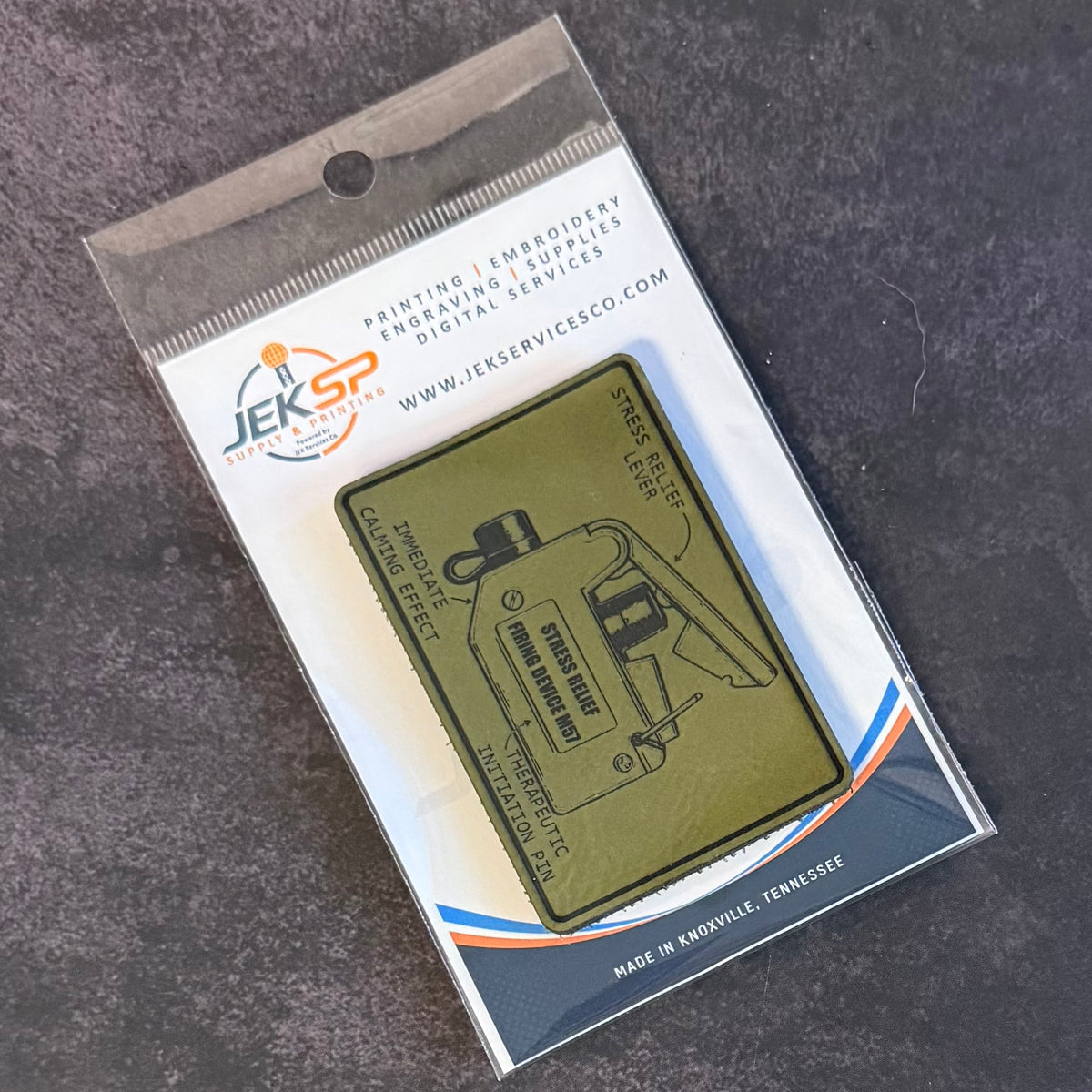 A JEK SP Service Company custom patch package containing an olive green rectangular patch with a black line-art engraving of a car, displayed on a dark textured background.