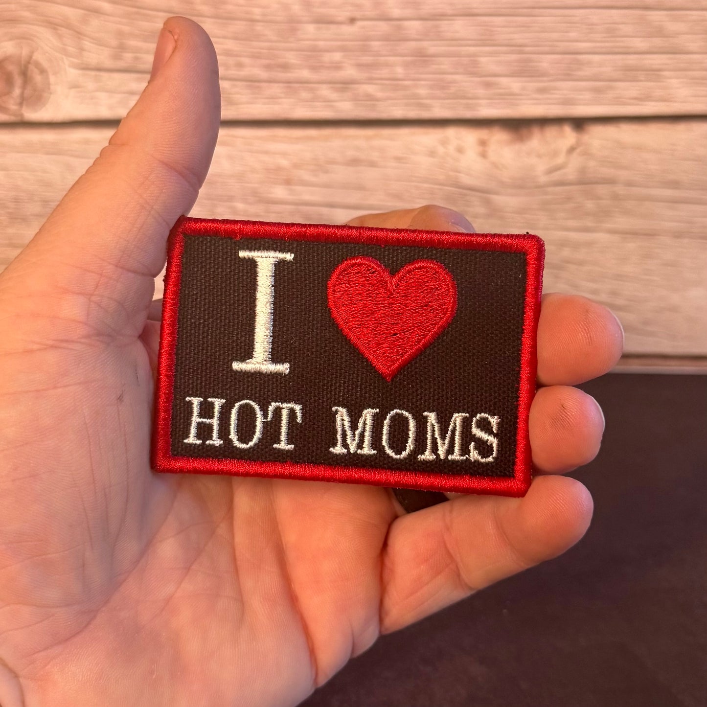A hand holding a rectangular embroidered patch that reads 'I [heart] HOT MOMS' in white and red on a dark brown background, with a light wood plank surface behind it.