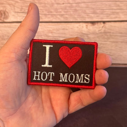 A hand holding a rectangular embroidered patch that reads 'I [heart] HOT MOMS' in white and red on a dark brown background, with a light wood plank surface behind it.
