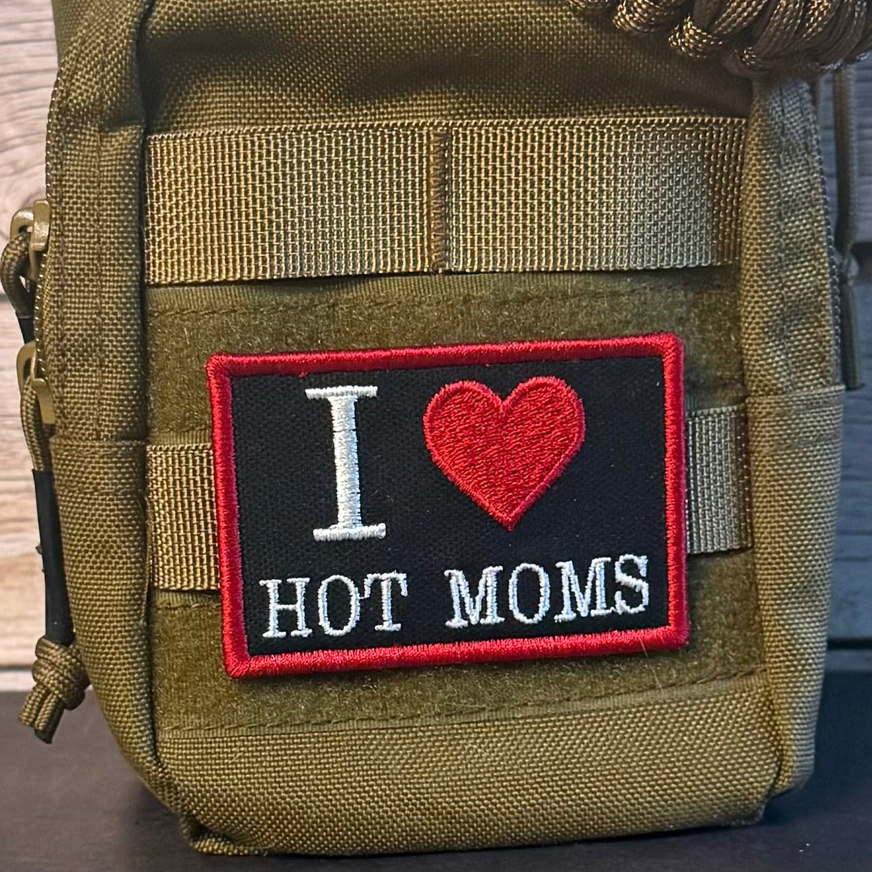 An olive drab tactical pouch with a front patch that reads 'I [heart] HOT MOMS' in white and red embroidery, shown against a rustic wood background.
