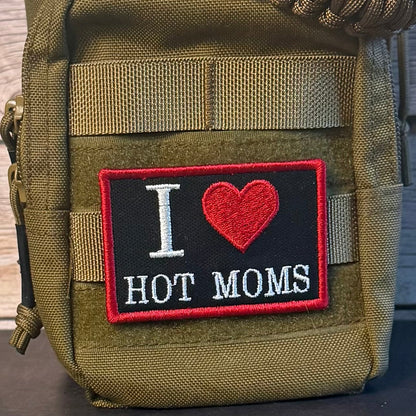 An olive drab tactical pouch with a front patch that reads 'I [heart] HOT MOMS' in white and red embroidery, shown against a rustic wood background.