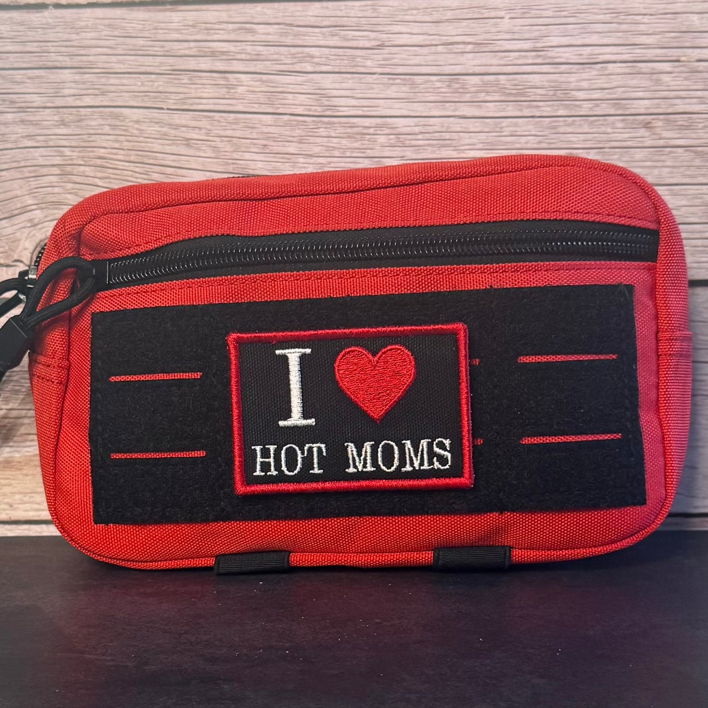 A red zippered pouch with a black front patch that reads 'I [heart] HOT MOMS' in white and red embroidery, placed against a light wood background.