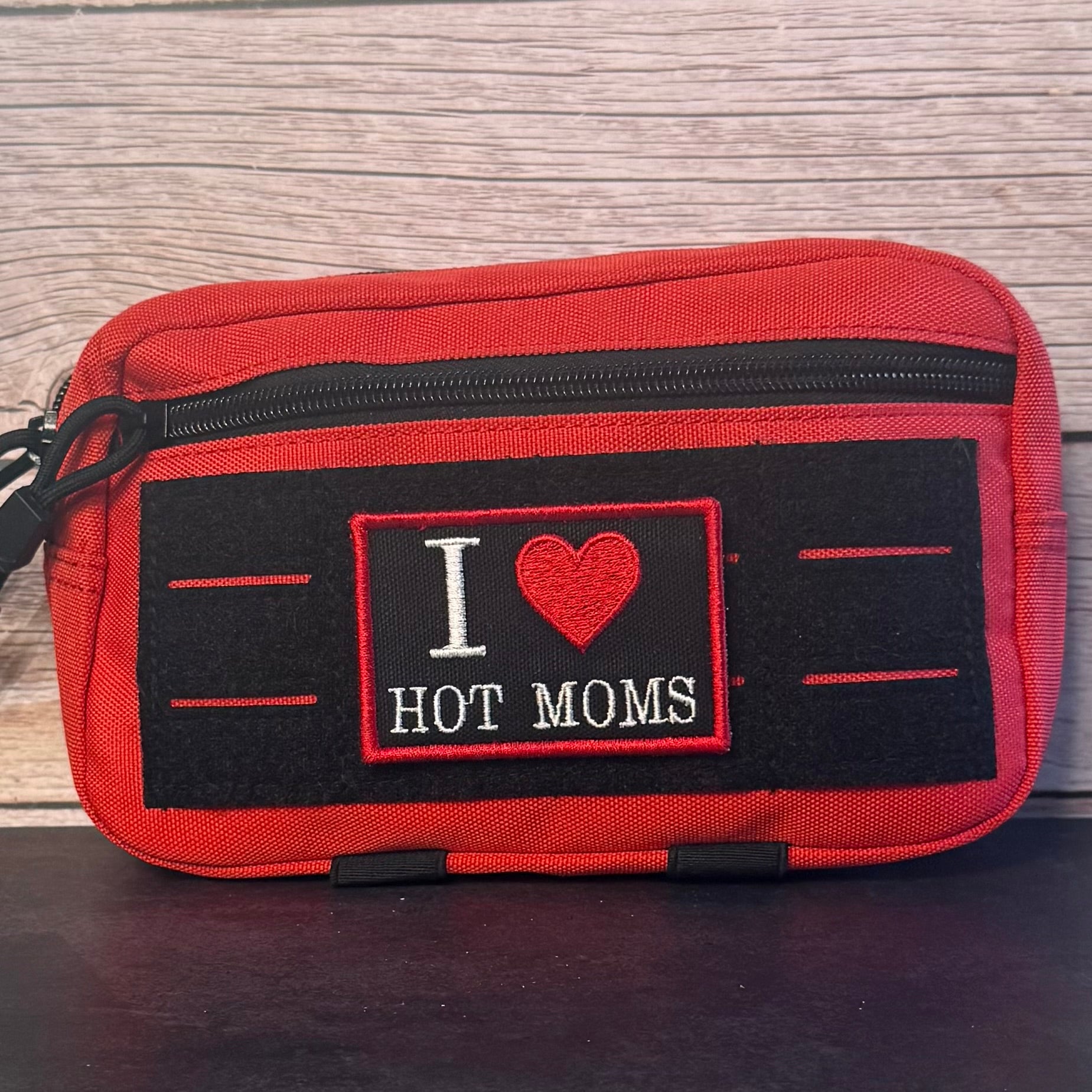 A red zippered pouch with a black front patch that reads 'I [heart] HOT MOMS' in white and red embroidery, placed against a light wood background.