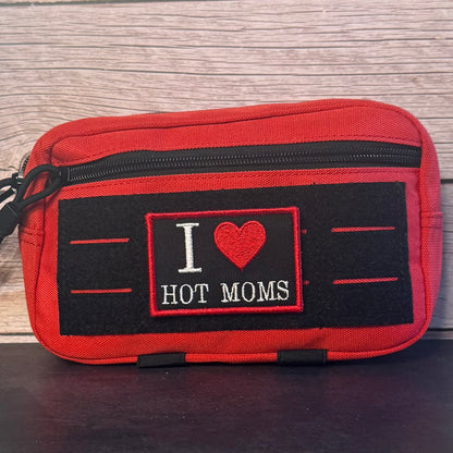 A red zippered pouch with a black front patch that reads 'I [heart] HOT MOMS' in white and red embroidery, placed against a light wood background.