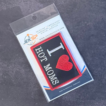An embroidered patch in its clear plastic packaging, featuring a black background with a red border, white text reading 'I HOT MOMS', and a red heart symbol.