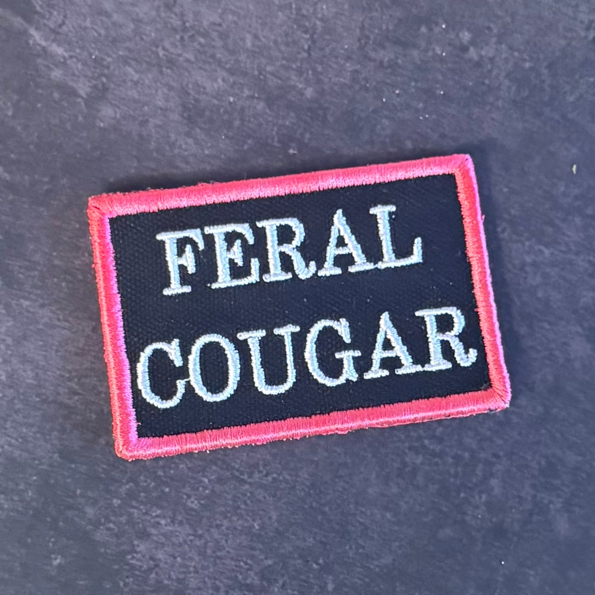 An embroidered rectangular patch with the words 'FERAL COUGAR' in white capital letters on a black background, bordered by a pink stitched edge, placed on a dark gray textured surface.