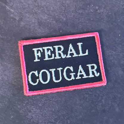 An embroidered rectangular patch with the words 'FERAL COUGAR' in white capital letters on a black background, bordered by a pink stitched edge, placed on a dark gray textured surface.