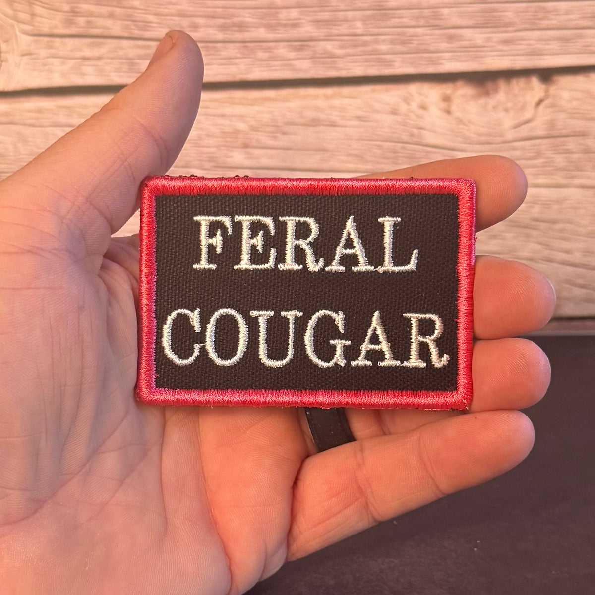 A hand holding a rectangular embroidered patch that reads 'FERAL COUGAR' in white lettering on a dark brown background with a red border, set against a light wood plank background.