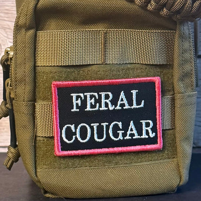 An olive green tactical pouch with a black and pink embroidered patch reading 'FERAL COUGAR' on the front, shown against a rustic wood background.