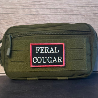 An olive green tactical pouch with a front zipper and a rectangular embroidered patch reading 'FERAL COUGAR' in white on a black background with a red border, placed against a light wood surface.
