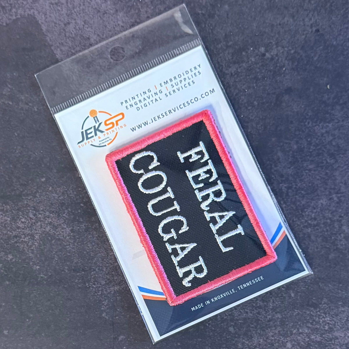 An embroidered patch in its retail packaging, featuring a black background with the words 'COUGAR' and 'FERAL' in white, bordered by pink and red.