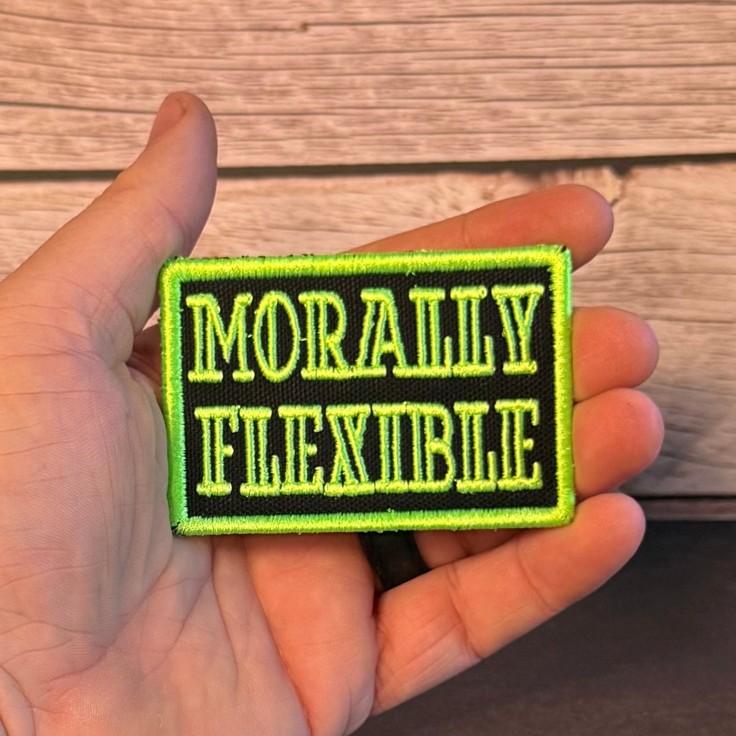 A hand holding a rectangular embroidered patch with the words 'MORALLY FLEXIBLE' in bright yellow-green lettering on a black background, set against a wood plank surface.