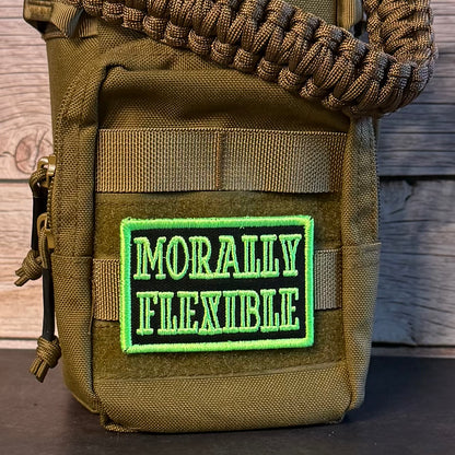 An olive drab tactical pouch with a woven paracord strap, featuring a rectangular embroidered patch that reads 'MORALLY FLEXIBLE' in green and black, set against a rustic wooden background.