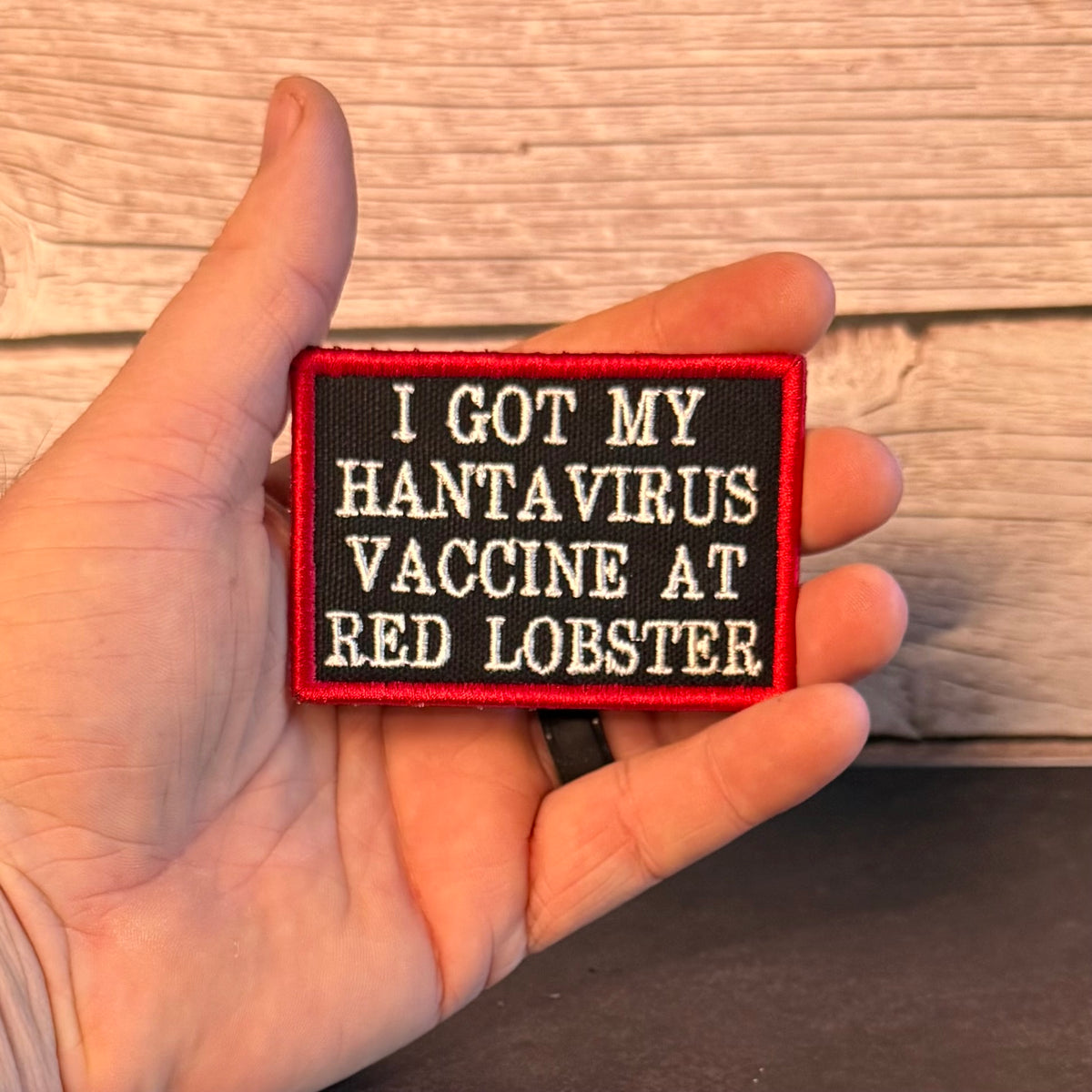 A hand holding a rectangular embroidered patch with a black background, red border, and white text that reads, 'I GOT MY HANTA VIRUS VACCINE AT RED LOBSTER.' The hand is positioned in front of a light wood plank background.