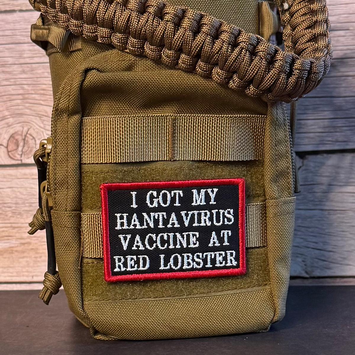 An olive green tactical pouch with a paracord strap, featuring a black and red embroidered patch that reads 'I GOT MY HANTAVIRUS VACCINE AT RED LOBSTER'.
