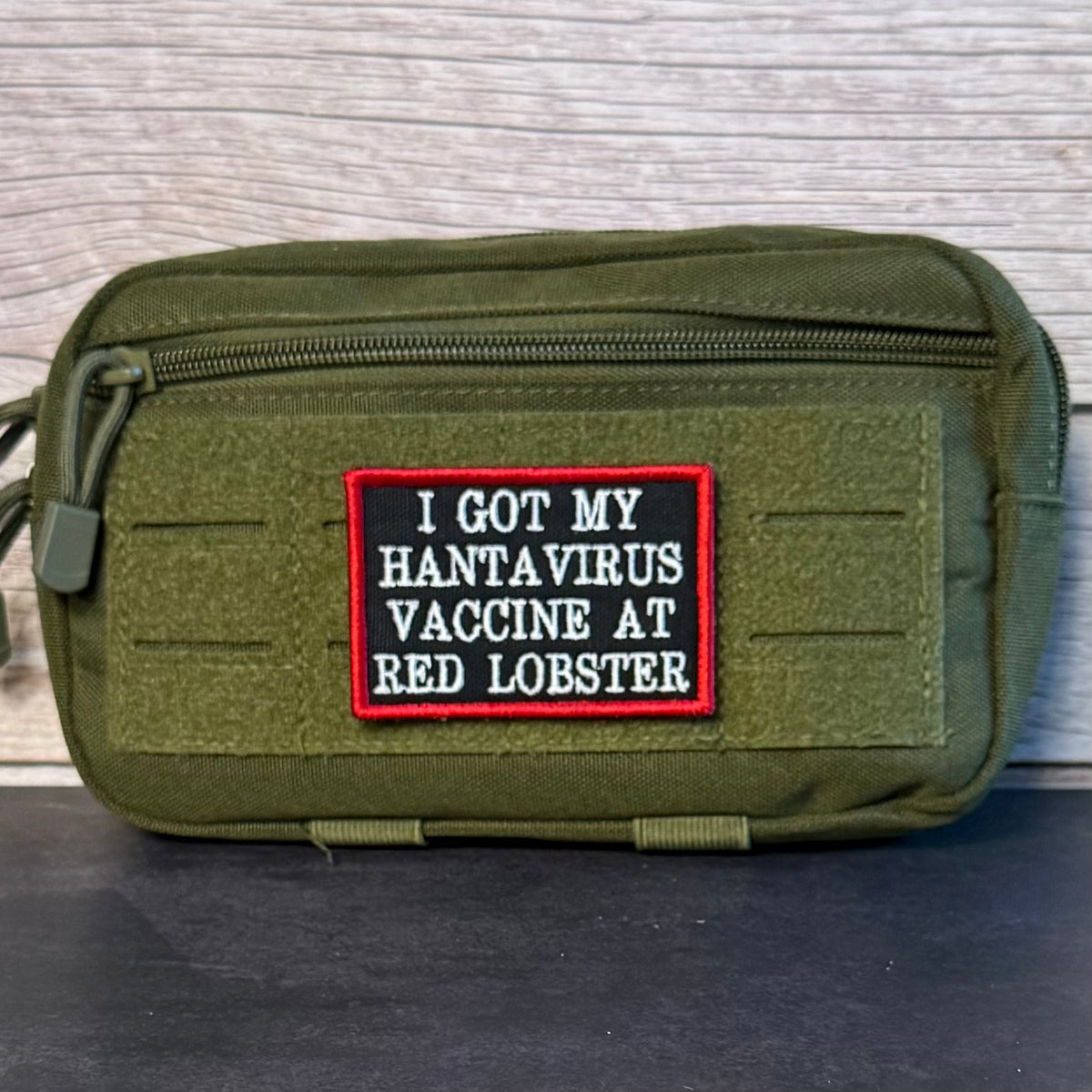 An olive green tactical pouch with a black and red embroidered patch that reads 'I GOT MY HANTAVIRUS VACCINE AT RED LOBSTER'.