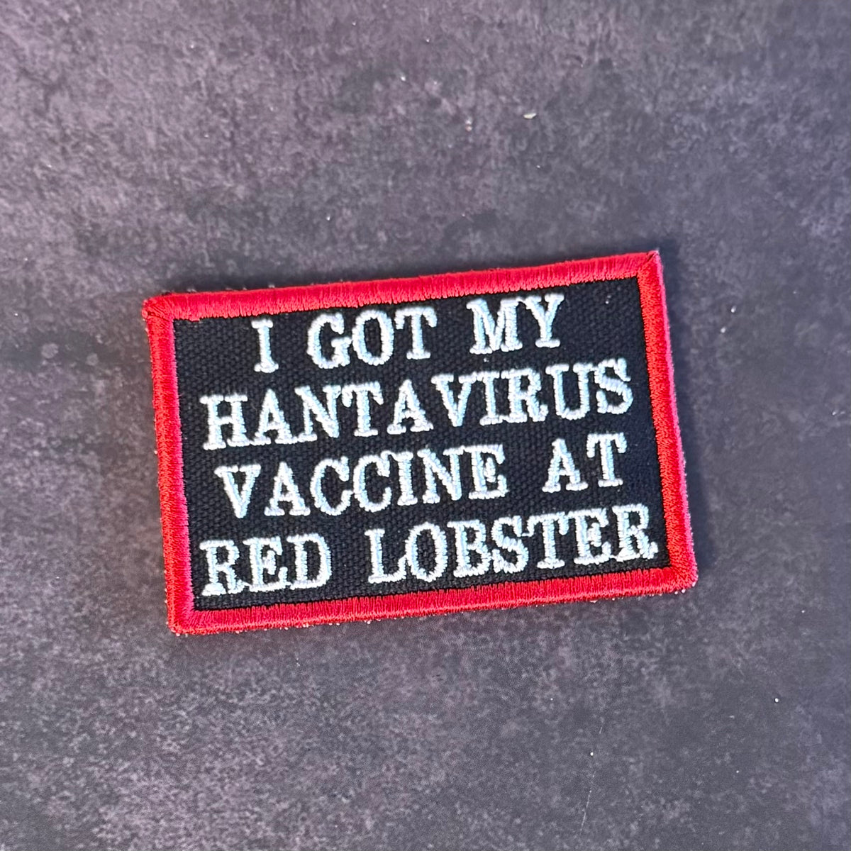 A rectangular embroidered patch with a black background, red border, and white text reading 'I GOT MY HANTAVIRUS VACCINE AT RED LOBSTER,' placed on a dark textured surface.