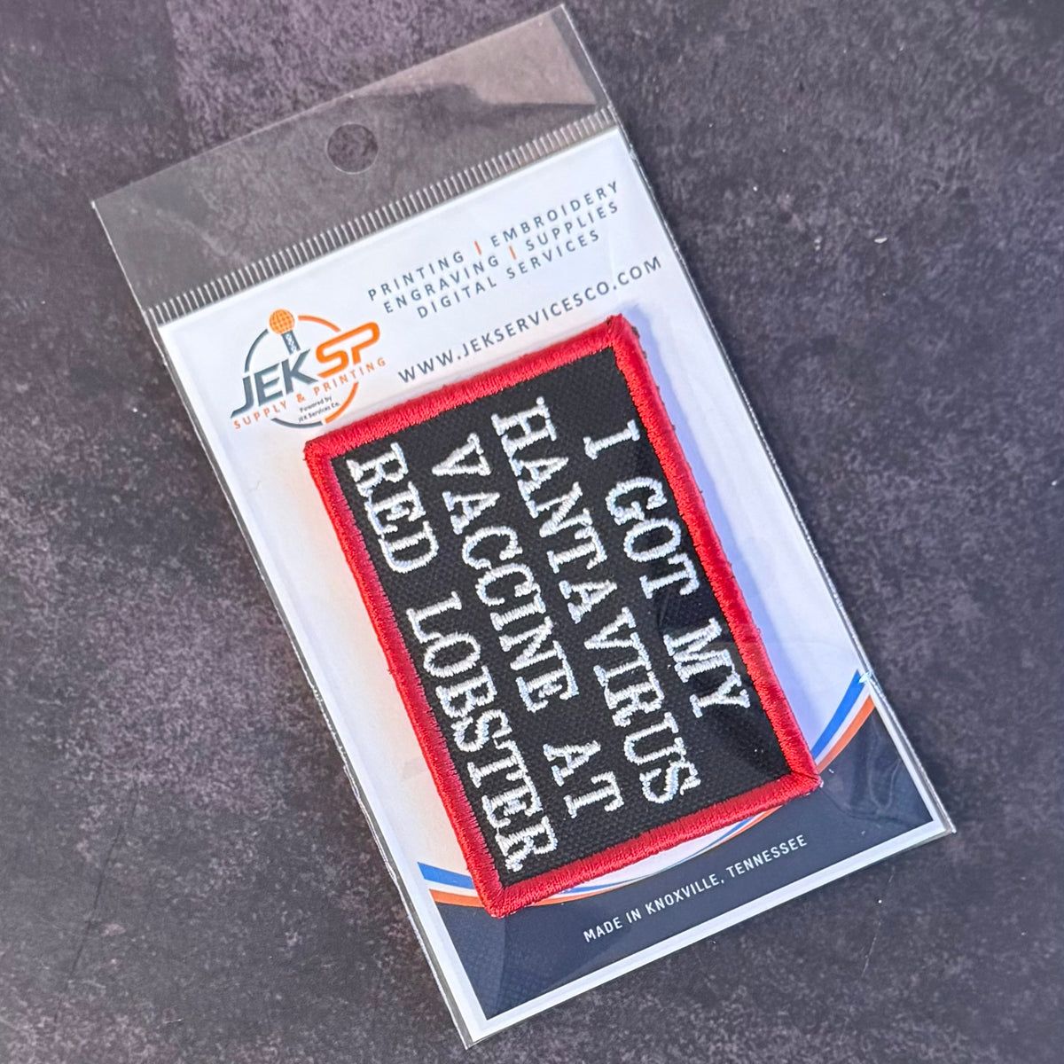 An embroidered patch in its clear plastic retail packaging, displayed on a dark textured surface. The patch features a black background with red stitched borders and white embroidered text reading 'I GOT MY HANTAVIRUS AT VACCINE RED LOBSTER'.