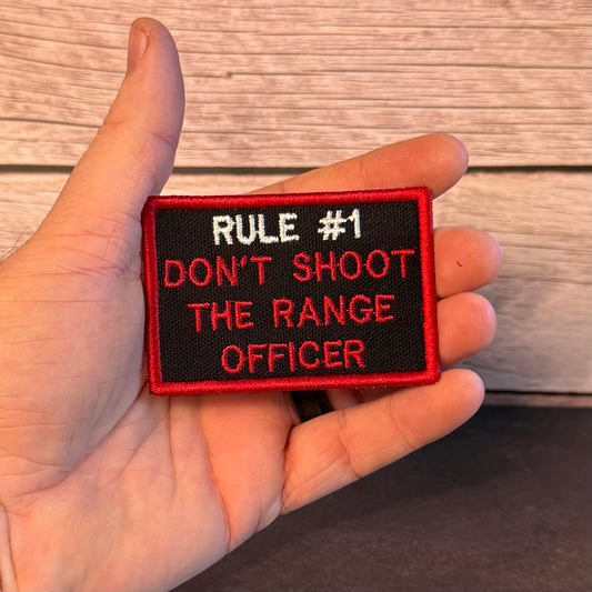 A hand holding a rectangular embroidered patch with the text 'RULE #1 DON'T SHOOT THE RANGE OFFICER' in white and red on a black background, set against a wood plank backdrop.