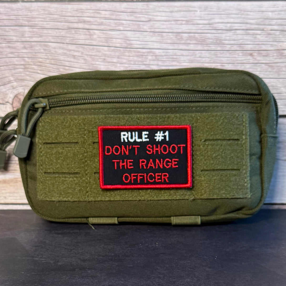 An olive green tactical pouch with a front zipper and a rectangular embroidered patch that reads 'RULE #1 DON'T SHOOT THE RANGE OFFICER' in red and white on a black background, set against a light wood plank backdrop.