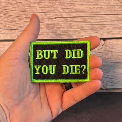 A hand holding a rectangular embroidered patch with the text 'BUT DID YOU DIE?' in bright green on a black background, set against a rustic wooden surface.