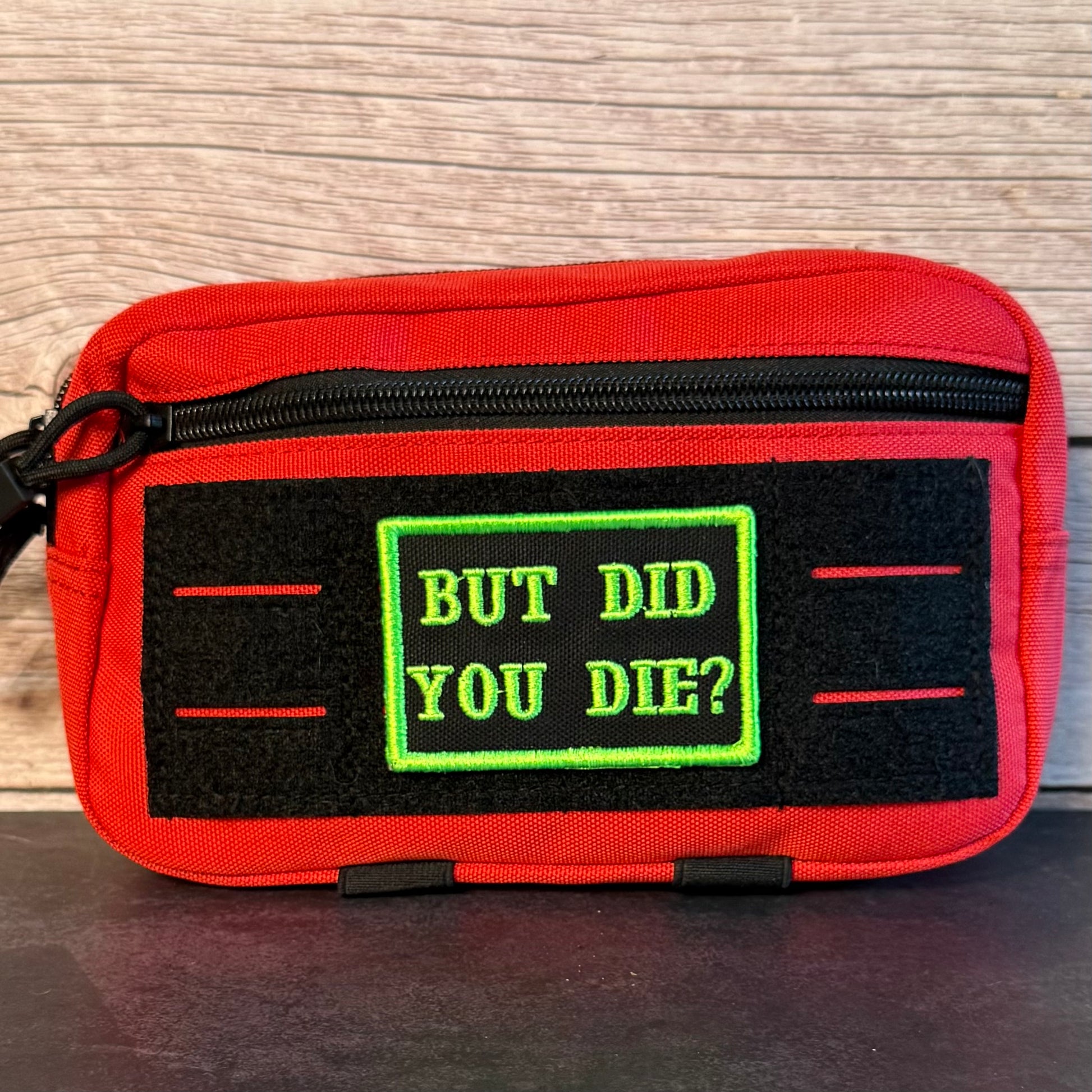 A red zippered pouch with a black velcro patch featuring the text 'BUT DID YOU DIE?' in green, set against a wood-grain background.