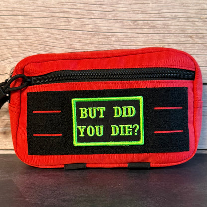 A red zippered pouch with a black velcro patch featuring the text 'BUT DID YOU DIE?' in green, set against a wood-grain background.