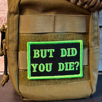 A close-up of a tactical pouch in olive drab and tan, featuring a black rectangular patch with bright green embroidered text that reads 'BUT DID YOU DIE?' and a bright green border. A brown paracord strap is threaded through the top zipper.