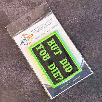 An embroidered patch reading 'BUT DID YOU DIE?' in bright neon green on a black background, sealed in clear plastic packaging with a printed backing card. The package is photographed on a dark textured surface.