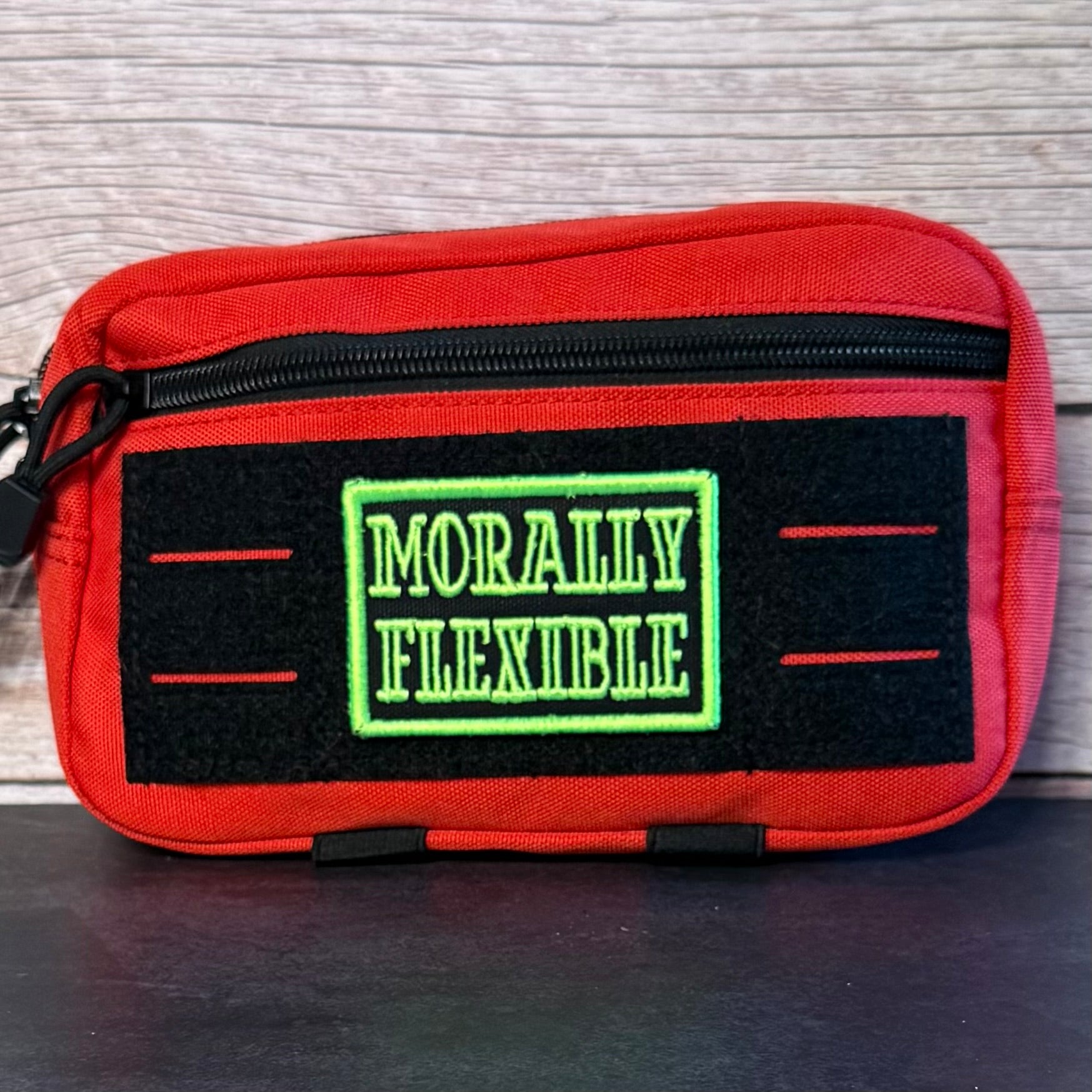 A red zippered pouch with a black front patch reading 'MORALLY FLEXIBLE' in green, photographed against a light wood background.