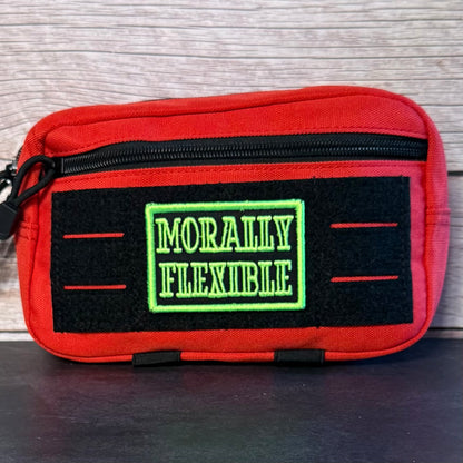 A red zippered pouch with a black front patch reading 'MORALLY FLEXIBLE' in green, photographed against a light wood background.