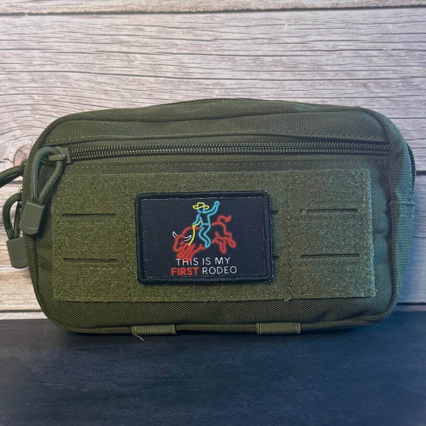 An olive green tactical pouch with a front hook-and-loop patch that reads 'THIS IS MY FIRST RODEO' with a cowboy and rider graphic, photographed against a wood plank background.