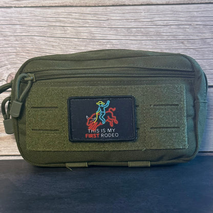 An olive green tactical pouch with a front hook-and-loop patch that reads 'THIS IS MY FIRST RODEO' with a cowboy and rider graphic, photographed against a wood plank background.