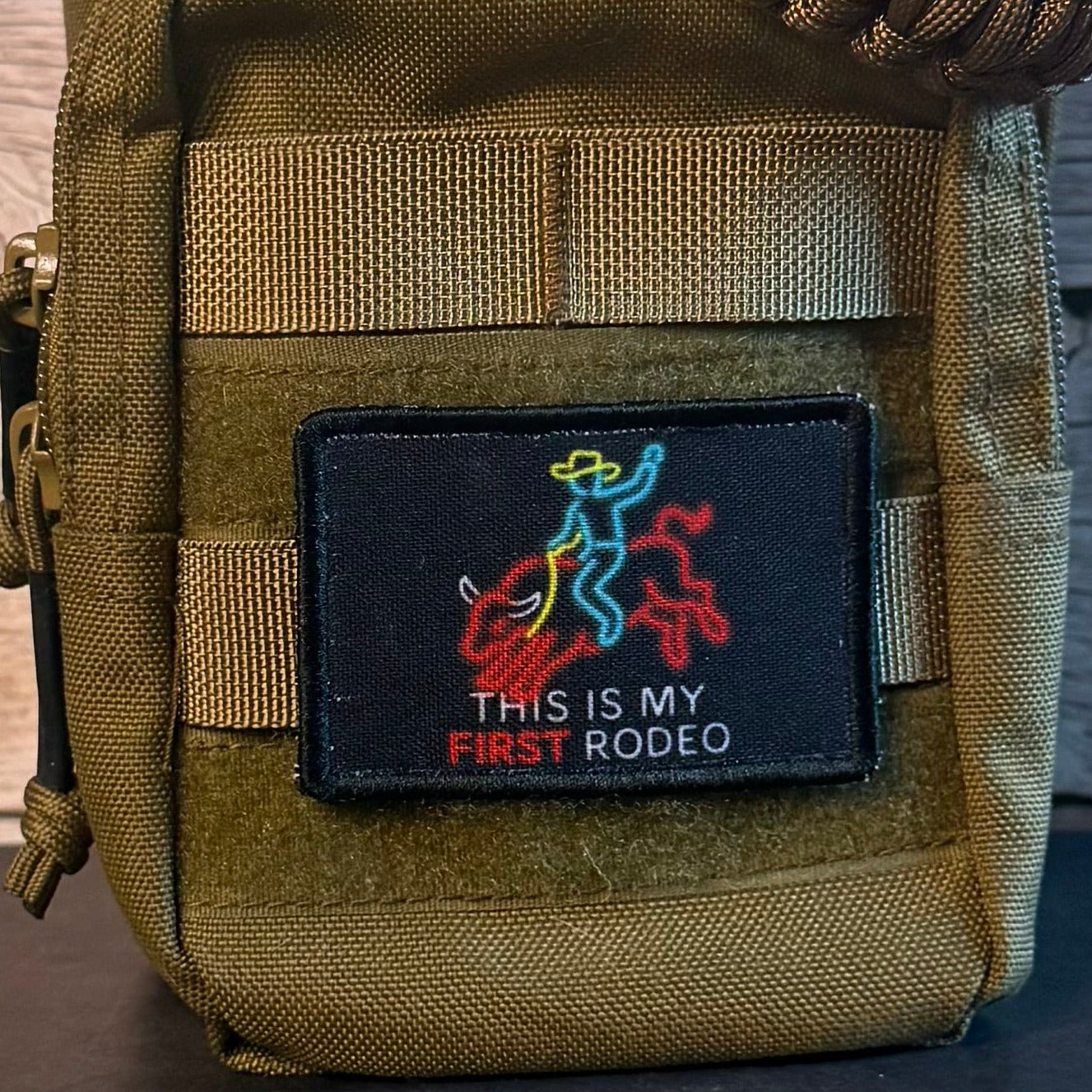 An olive green tactical pouch with a black rectangular patch that reads 'THIS IS MY FIRST RODEO' with a colorful cowboy and rider graphic, shown against a dark wood background.