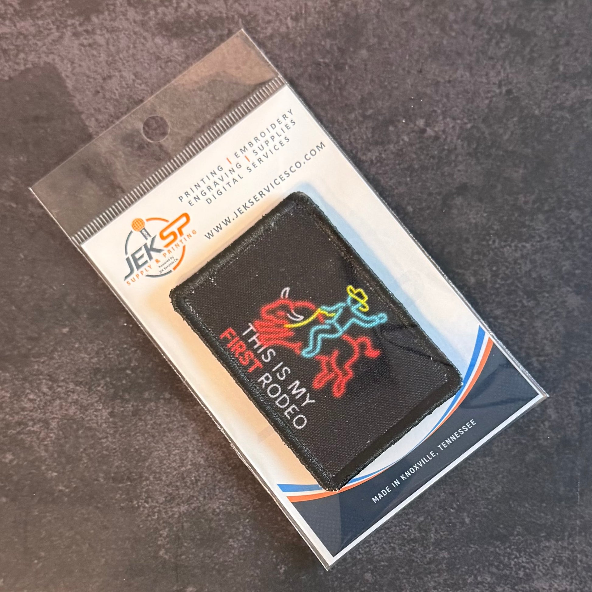 A black rectangular embroidered patch with a colorful neon-style horse graphic and the text 'THIS IS MY FIRST RODEO' in white, still inside clear retail packaging on a dark textured surface.