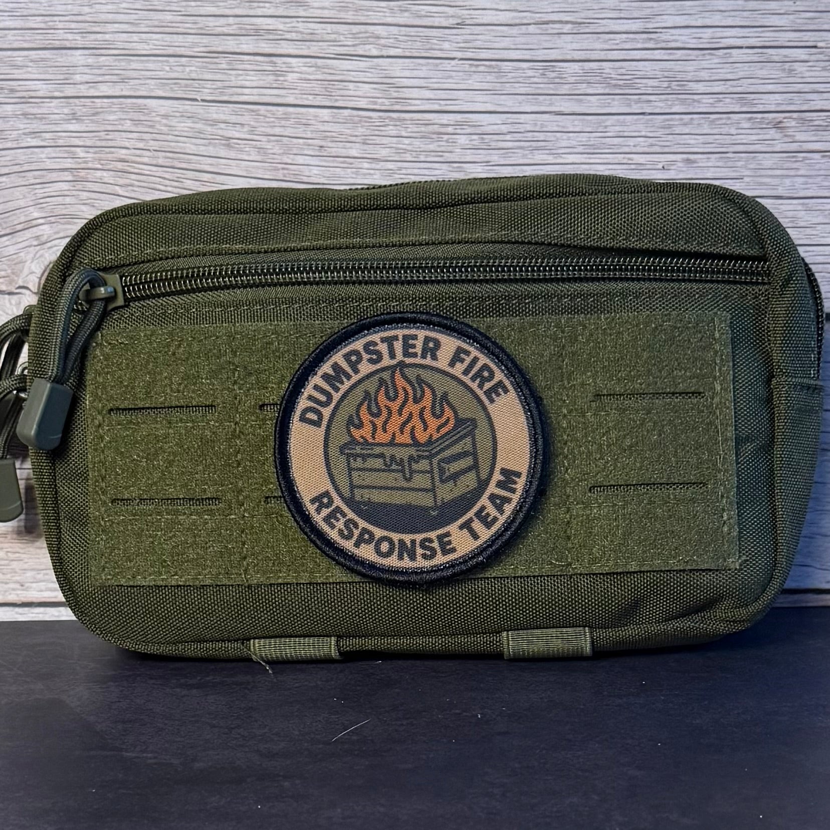 An olive green tactical pouch with a circular 'Dumpster Fire Response Team' patch on the front, set against a gray wood-grain background.