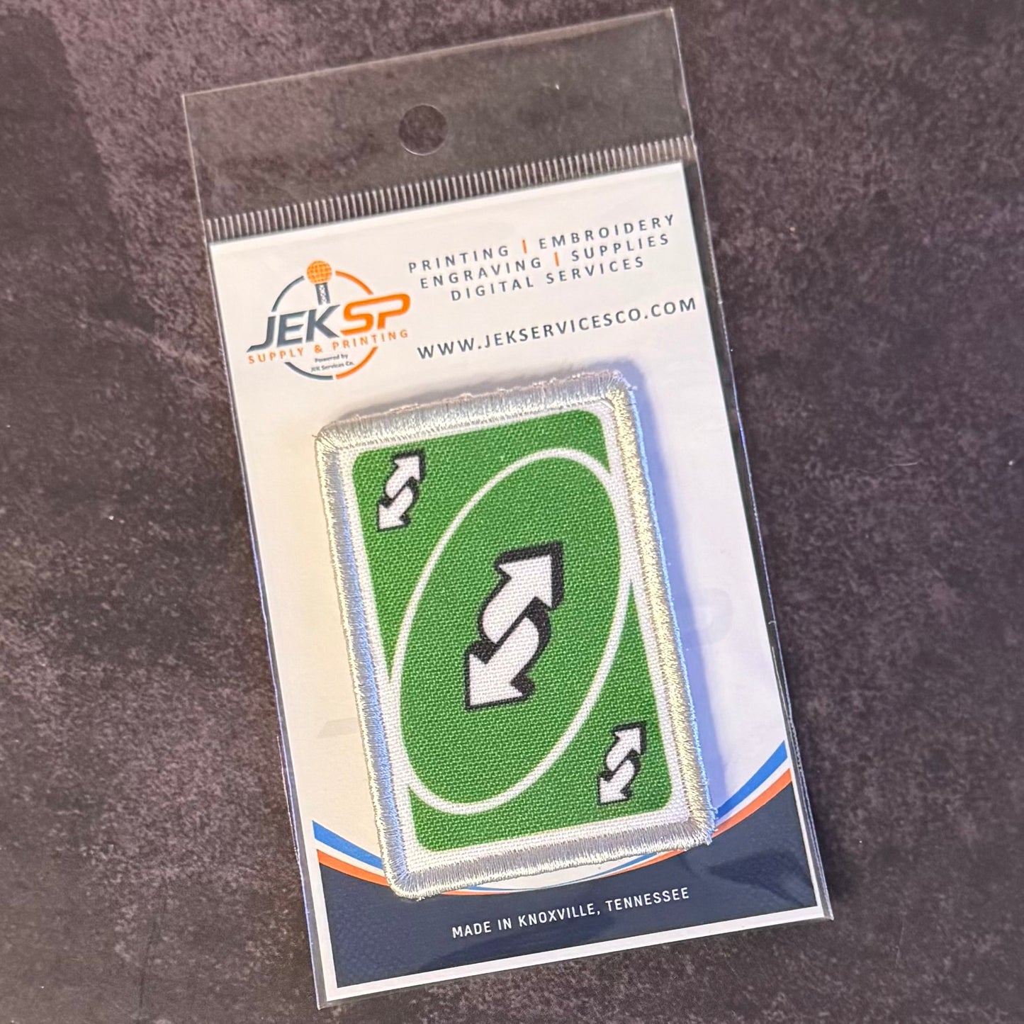 A green rectangular patch with a white border and three white arrow symbols, sealed in a clear plastic package with a printed backing card. The backing card shows the JEKSP Supplies Services logo and text.