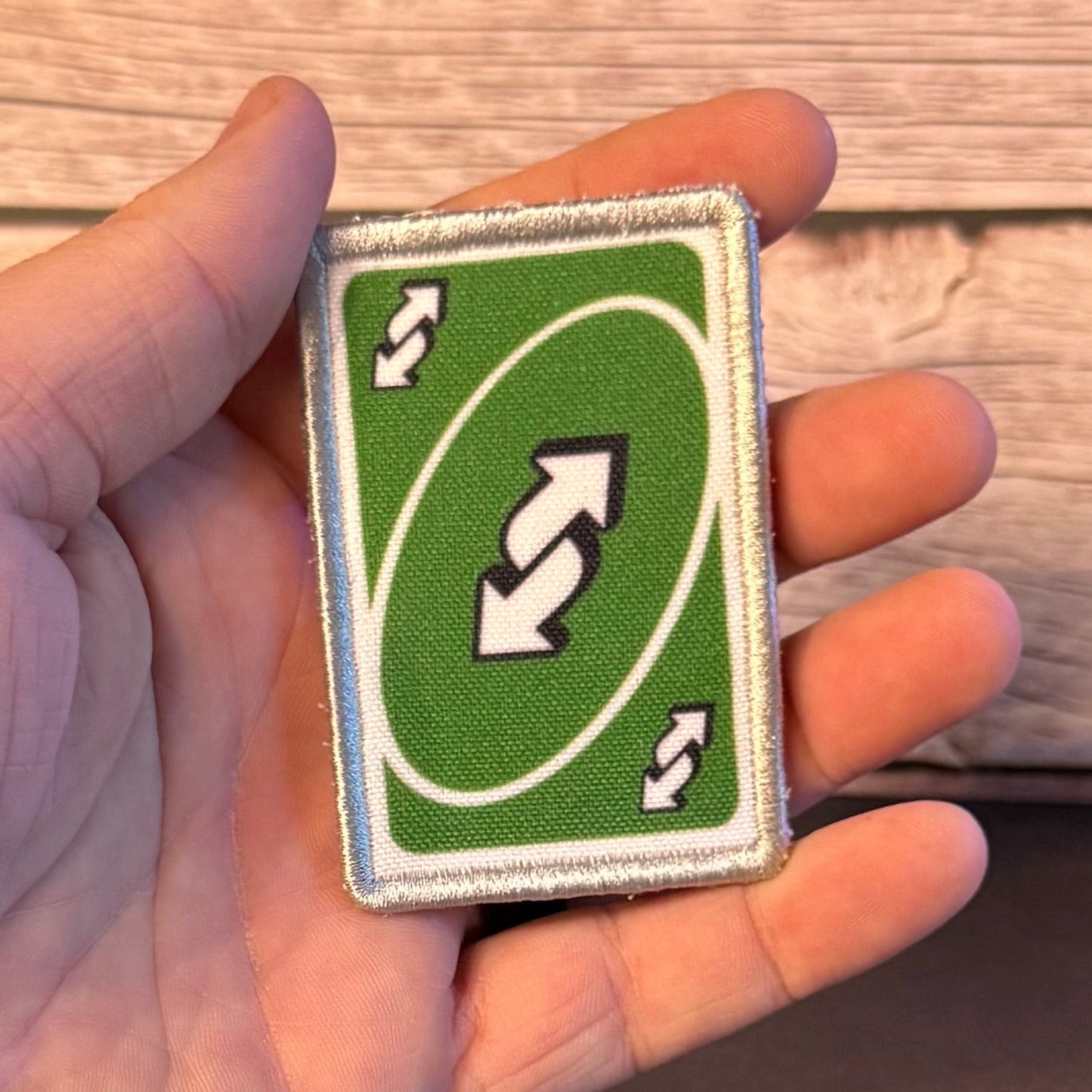A hand holding a small embroidered patch shaped like a green playing card with white border, a double-arrow symbol in the center, and small arrow icons in the corners, set against a wooden background.