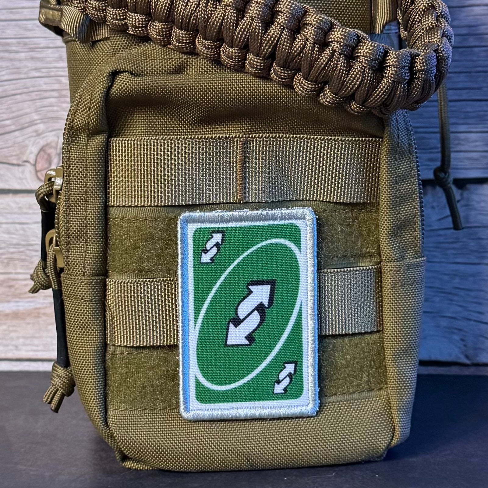 A close-up of a tactical pouch in olive drab green nylon with a woven brown paracord loop at the top and a green rectangular patch featuring a white double-arrow loop symbol, set against a rustic wood background.