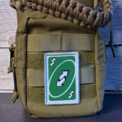 A close-up of a tactical pouch in olive drab green nylon with a woven brown paracord loop at the top and a green rectangular patch featuring a white double-arrow loop symbol, set against a rustic wood background.