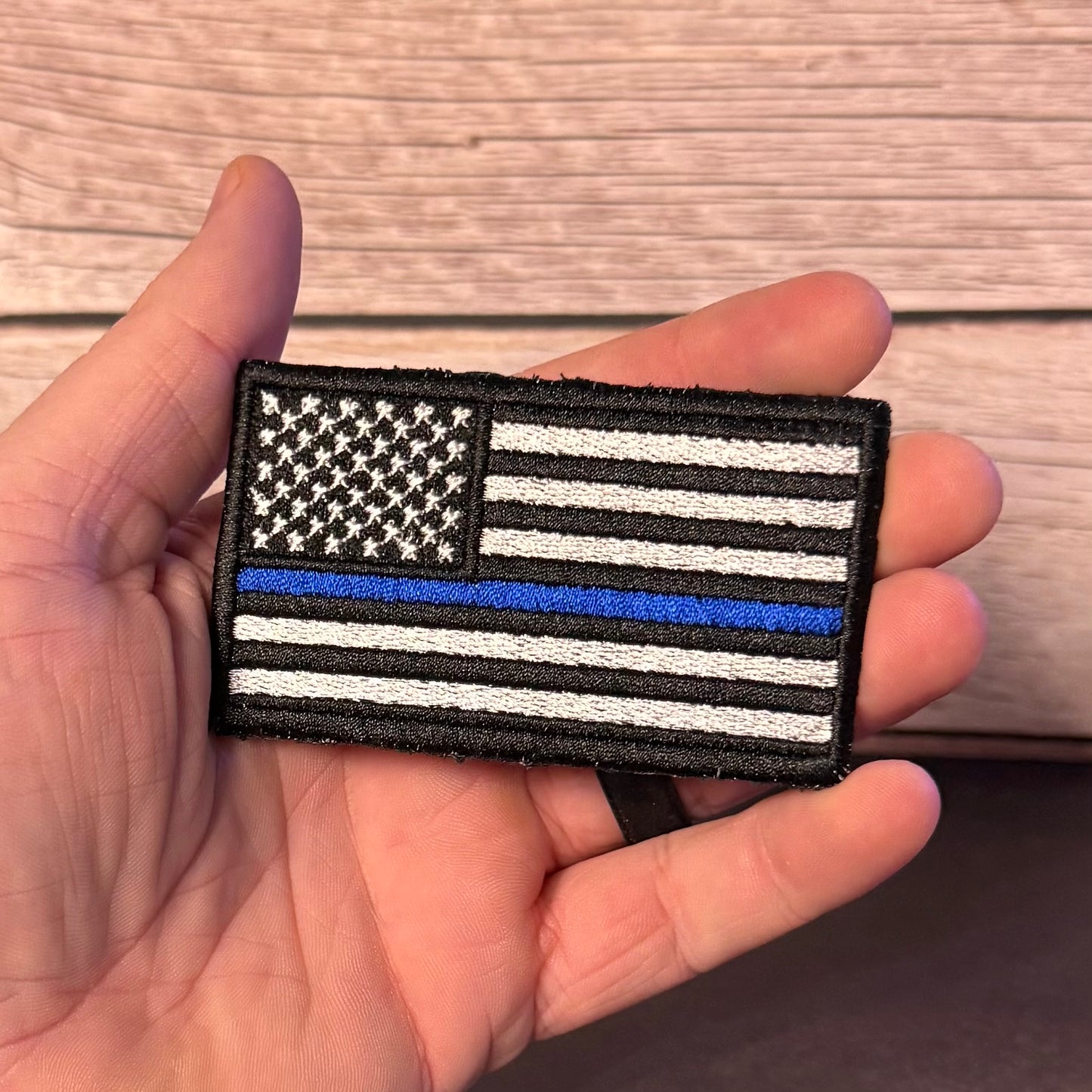 A hand holding a rectangular embroidered patch featuring a black-and-white American flag with one blue stripe, shown against a wood plank background.