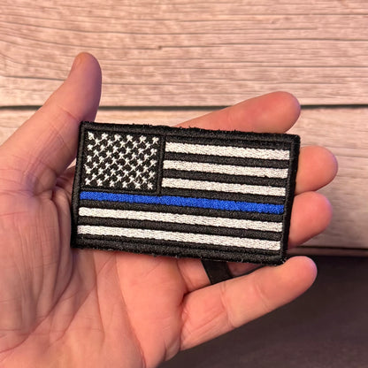 A hand holding a rectangular embroidered patch featuring a black-and-white American flag with one blue stripe, shown against a wood plank background.