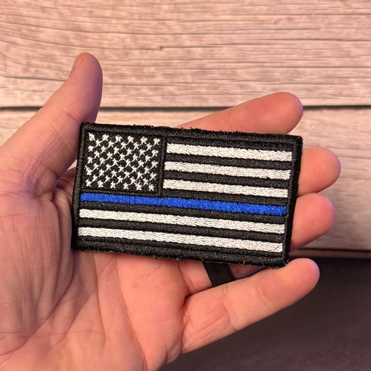 A hand holding a rectangular embroidered patch featuring a black-and-white American flag with one blue stripe, shown against a wood plank background.