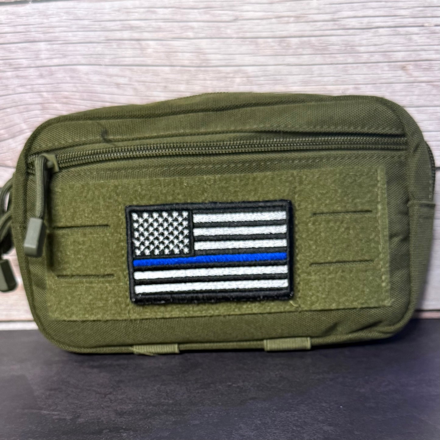 An olive green tactical pouch with a zippered top and a black, white, and blue embroidered American flag patch on the front, set against a light wood background.