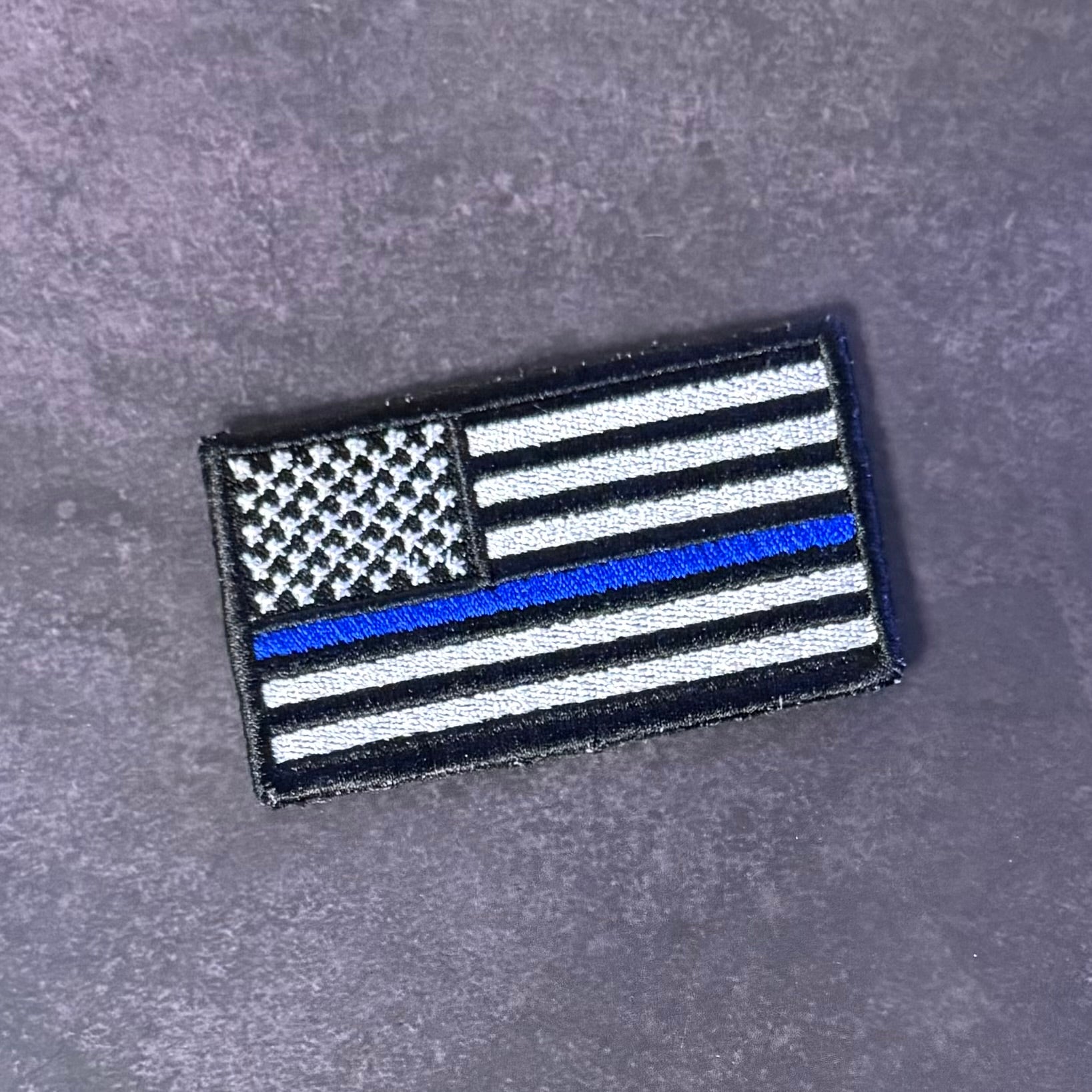 A rectangular embroidered patch featuring a black and white American flag design with a single blue stripe, placed on a textured gray surface.
