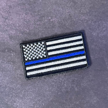 A rectangular embroidered patch featuring a black and white American flag design with a single blue stripe, placed on a textured gray surface.