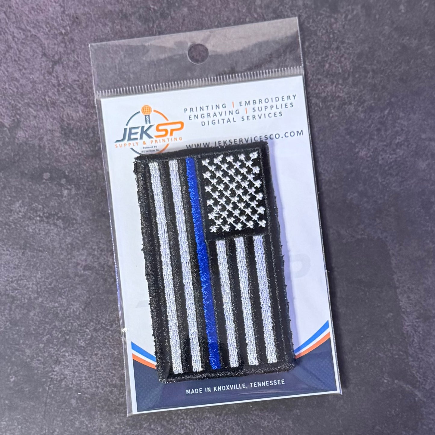 A 'Thin Blue Line' American flag embroidered patch in black, white, and blue, packaged on a white backing card with the JEKSP Printing logo, set against a dark gray textured background.
