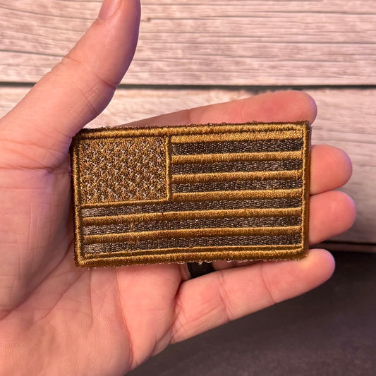 A close-up of a hand holding a rectangular embroidered American flag patch in coyote brown and tan brown thread, with a wood plank background.