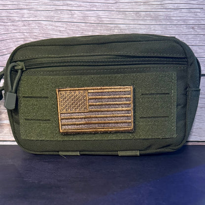 An olive green tactical pouch with a front zipper and a brown embroidered American flag patch on the front, set against a gray wood plank background.
